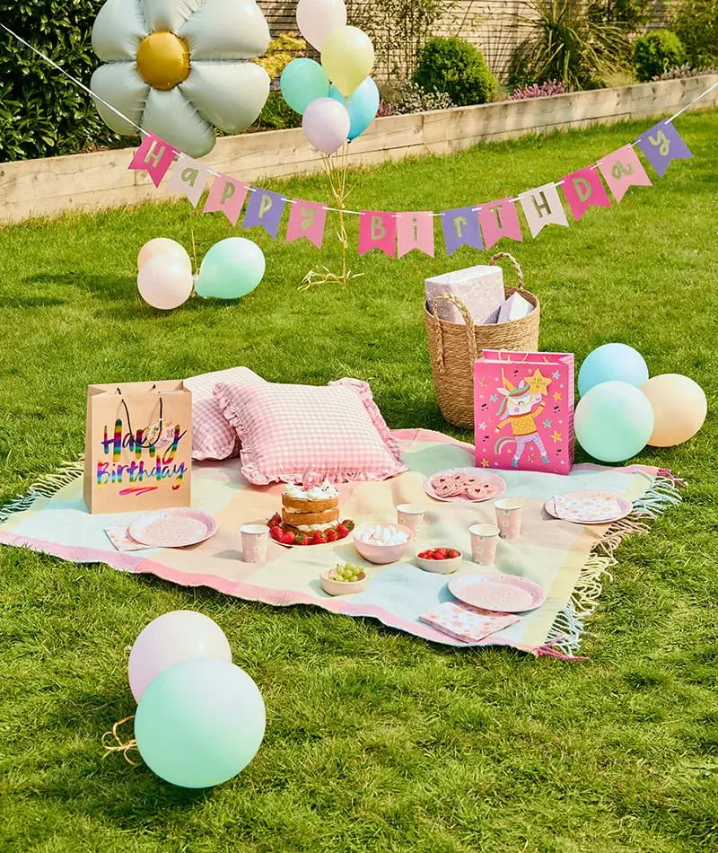 Pastel birthday picnic setup with a birthday cake, balloons, gift bags, pink tableware, cushions, and a Happy Birthday banner on a garden lawn.